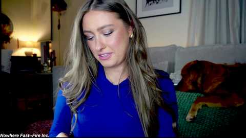 Video of a young woman with long, wavy, light brown hair, wearing a bright blue top, sitting in a cozy living room with a beige couch and a large dog lying on a green blanket.