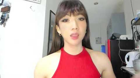 Video of a young Asian woman with long brown hair, wearing a red halter top, making a playful face, in a cluttered, modern kitchen.