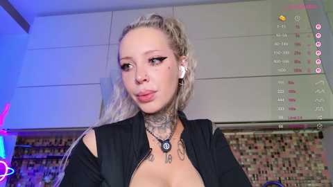 Video of a blonde, tattooed woman with a high ponytail, wearing a black top, standing in a modern kitchen with a calendar and colorful lights in the background.