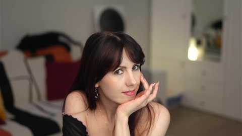 Video of a young Caucasian woman with long dark hair, light skin, and a slender build, smiling and resting her chin on her hand in a cozy, dimly-lit room.