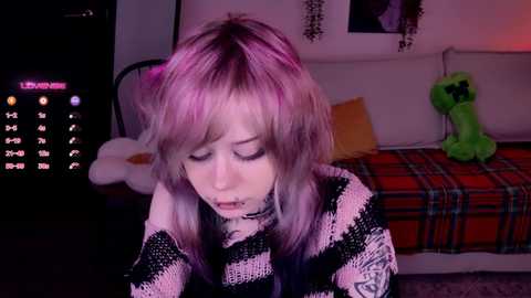 Video of a pale-skinned woman with shoulder-length, multi-colored hair, wearing a black and white striped sweater, seated in a dimly lit room with a TV displaying \"Live Stream\" and a green plush dinosaur toy.