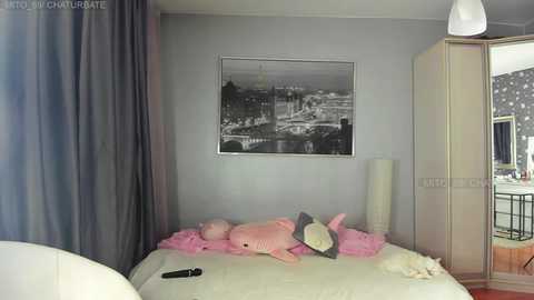 Video of a modern, minimalist bedroom with a large gray wall, a black-and-white cityscape print, a plush pink shark toy on a white bed, and a gray curtain.