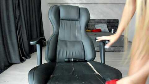 Video of a black leather gaming chair in a modern, minimalist room with beige walls, grey couch, and black curtains. A woman with long blonde hair is adjusting the chair's armrests.
