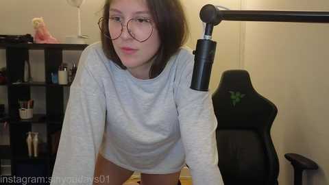 Video of a young woman with glasses, wearing a gray sweater, leaning over a black gaming chair. Background includes a shelf with various items, beige walls, and a lamp.