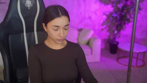 Video of a woman with long dark hair in a black gaming chair, wearing a tight black long-sleeve top, in a softly lit room with purple ambient lighting.