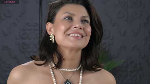 Video of a smiling, light-skinned woman with shoulder-length dark hair, wearing a pearl necklace and gold earrings, against a dark, floral-patterned background.