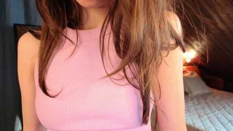 Video of a young woman with long, brown hair, wearing a sleeveless, ribbed pink top, indoors with a bed and lamp in the background.