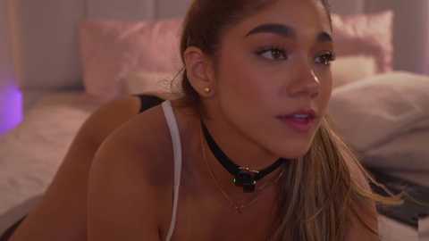 Video of a young woman with light brown skin, brown hair, wearing a white tank top and black choker, lying on a bed with soft pink pillows and sheets.
