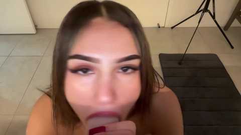 A video of a topless woman with long, straight brown hair, performing oral sex on a man. She has fair skin, full lips, and dark eyebrows. The setting is a beige-tiled bathroom with a black rug and a tripod.