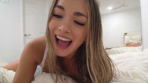 Video of a young woman with long, blonde hair, light skin, and closed eyes, smiling widely in a bright bedroom with white walls, a bed, and a mirror.