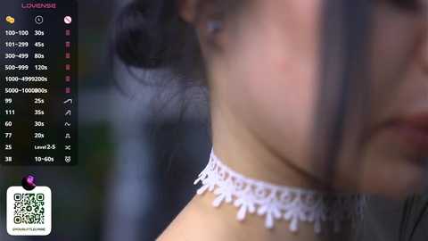 Video of a woman's face with long, black hair, wearing a white lace choker, in a blurred background. QR code and digital interface with video controls on the left.