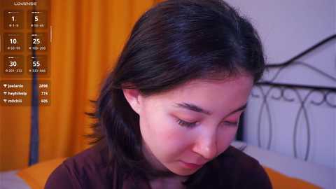 Video of a young Asian woman with long, dark hair, wearing a dark top, seated on a bed with white sheets and orange curtains in the background.
