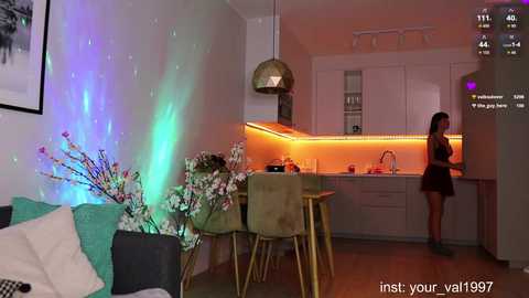 Video of a modern, dimly-lit living room with vibrant LED lights casting a multi-colored glow, featuring a woman in a dark dress standing near a sleek kitchen with white cabinets.