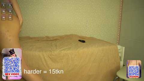 A video showing a nude woman with dark skin, medium build, and small breasts, standing near a beige bed with a brown quilt. The room has textured green walls.