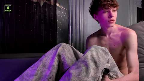 Video of a young, shirtless, fair-skinned male with short brown hair, covered in a grey blanket, sitting on a bed, with dim, purple-lit room background.