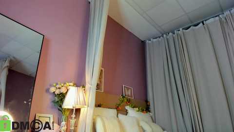 Video of a cozy bedroom with lavender walls, a large mirror, white curtains, and a white bed with pillows. A table holds a lamp, flowers, and framed pictures. The room has a soft, inviting ambiance.