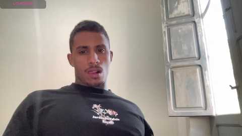 A young, light-skinned African American man with short hair, wearing a black shirt with a colorful graphic, stands in a dimly lit room with a window and a door visible in the background.