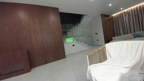 Video of a modern, minimalist living room with a wooden wall, white couch, and beige curtains; visible stairs and glass railing in the background.