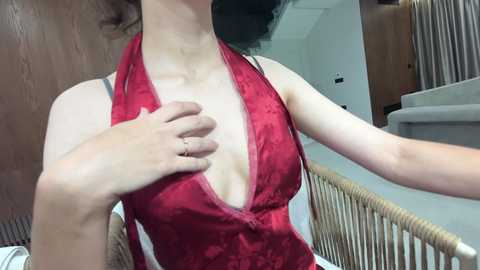 Video of a light-skinned woman in a revealing red silk halter dress, with her right hand over her left breast, standing in a modern, minimalistic bathroom with a wooden door and white sink.