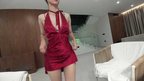 Video of a woman in a red halterneck dress, standing indoors with modern wooden walls, white lounge chairs, and a staircase in the background.