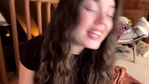 A video captures a young woman with long, wavy brown hair, wearing a black shirt, laughing joyfully with closed eyes. She's in a cozy, wooden-paneled room with a warm ambiance.