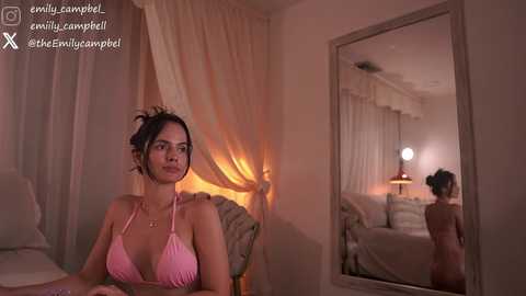 A video of a woman in a pink bikini sitting in a dimly lit bedroom, with a large mirror reflecting her image and the room's cozy ambiance.
