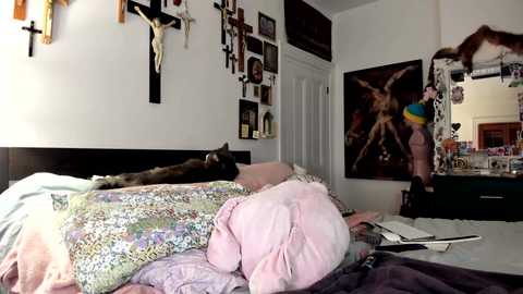 Video of a cluttered bedroom with a black crucifix, a cat on a bed, colorful blankets, a yellow hat, and religious wall art.