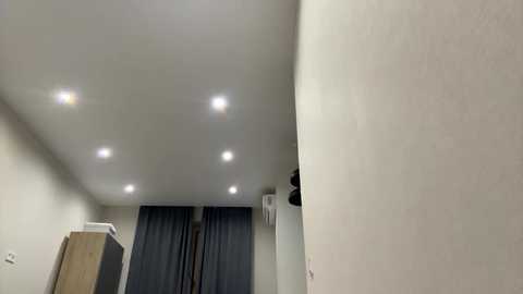 Video of a modern, minimalist room with a smooth, light beige wall and a large window covered by dark gray curtains. The ceiling has recessed lighting. The room has a white refrigerator and a wooden cabinet on the left.