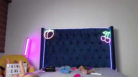 A video of a modern, dark blue, tufted headboard with neon pink and green lights. A person in a yellow shirt and glasses stands in front, holding a sign reading \"Gina Cali.\" The room is cluttered with toys and colorful items.