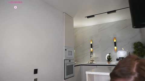 Video of a modern kitchen with light grey marble backsplash, sleek white cabinets, built-in ovens, and two wall-mounted lamps with black and gold bases.