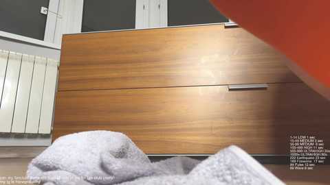 Video of a modern, wooden dresser with a polished finish and silver handles, set against a gray wall with a white radiator. A light gray towel is draped over the dresser's top left corner.