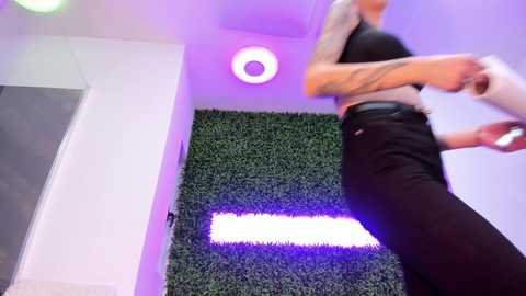 Video of a muscular, tattooed woman in a tight black top and pants, dancing energetically against a green faux-grass wall, illuminated by purple and white lights.