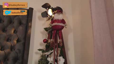 Video of a festive Christmas decoration featuring a plush Santa Claus doll with a red suit, white fur trim, and a black hat, standing on a green garland with white flowers and red ornaments.