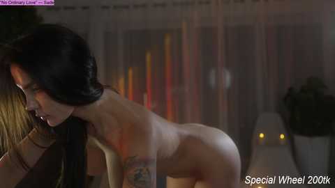 A video of a nude woman with long black hair and tattoos, bending forward, in a dimly lit room with blurred, colorful lights in the background.