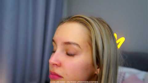 Video of a young woman with fair skin and blonde hair, wearing pink lipstick, resting her head on a grey pillow in a bedroom.