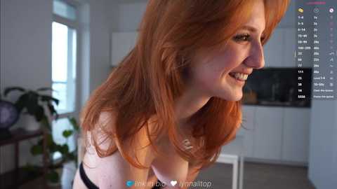 A video of a smiling, fair-skinned woman with long, wavy red hair, wearing a strapless top, in a modern, well-lit kitchen.