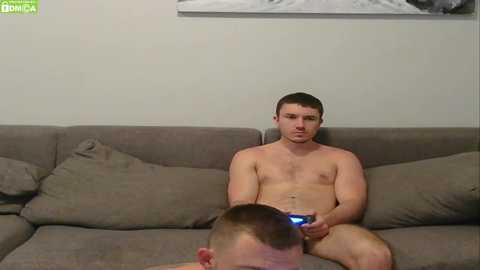 Media: Video of a shirtless, muscular, light-skinned man with short brown hair, playing a video game on a couch, with another man's head visible in the foreground.