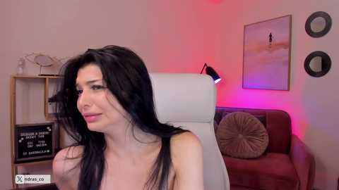 Video of a fair-skinned woman with long black hair, wearing a black tank top, seated in a white chair in a cozy, dimly lit room with red lighting, a brown couch, and wall art.