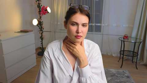 Video of a young woman with light skin, brown hair, wearing white robe, resting her hand on her neck, standing in a softly lit room with white walls, a table, and a tall flower vase.