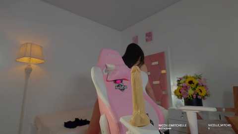 Video of a young woman with straight black hair, wearing a yellow dress, seated in a pink gaming chair, surrounded by white walls, a lamp, and a flower arrangement.