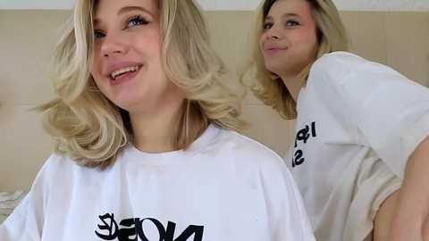 Video of a blonde Caucasian woman smiling, wearing a white T-shirt with black text, standing in a beige-tiled room with a mirror reflection.