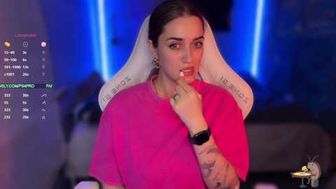 Video of a young woman with light skin and dark hair in a pink t-shirt, sitting in a white gaming chair, with blue lighting and a live-streaming setup in the background.