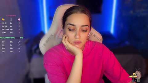 Media: A video of a young woman with light skin and brown hair in a pink sweater, resting her head on her hand, in a dimly lit room with blue LED lighting.