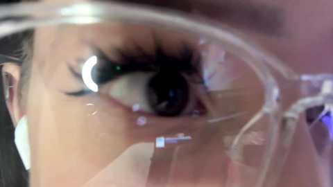 Video of a close-up, partially obscured face with a clear plastic covering over the mouth and nose, highlighting the eyes' intense makeup with thick, black eyeliner and mascara.