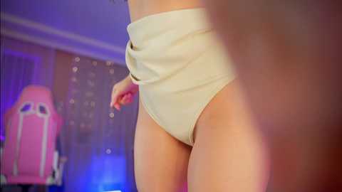 A video of a fair-skinned woman wearing beige high-waisted panties in a dimly lit room with a pink gaming chair and string lights in the background.