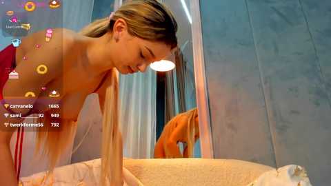 Video of a blonde woman with fair skin, topless, leaning over a bed, wearing a sheer pink dress. She gazes at her reflection in a mirror, surrounded by soft, warm lighting.