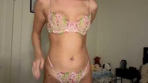 A video of a slender, light-skinned woman with a flat stomach wearing a pastel pink floral bra and panties, standing in a modestly furnished bedroom with a white door and stuffed animals in the background.