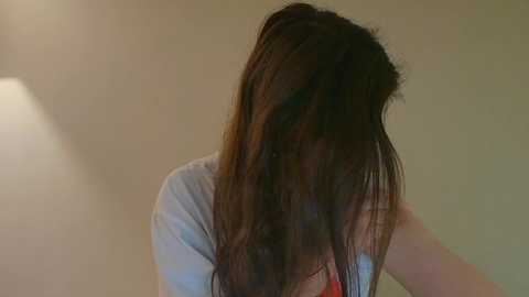 Video of a woman with long, dark brown hair, wearing a light gray T-shirt, leaning forward with her head down, against a beige background.