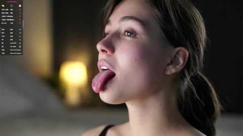 Media: Video of a fair-skinned young woman with brown hair, sticking out her tongue, wearing a black top. Background is blurred with a yellow light source.