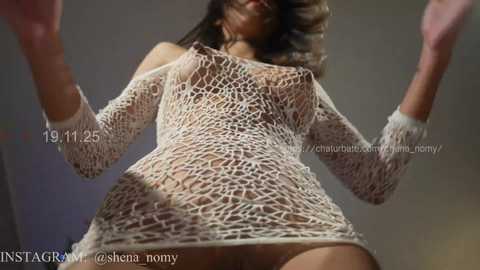 Video of a woman with light brown skin, wearing a white, fishnet, off-shoulder dress, revealing her breasts and pubic hair. She is in a low-angle, close-up shot, indoors with blurred background.